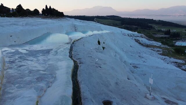 A drone flies smoothly forward, tracking over the vast white travertine terraces of Pamukkale, Turkey. Soft morning dawn light glows across the organic limestone formations and natural thermal pools, 