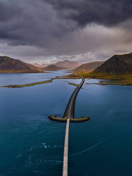 Aerial view of a road stretching across the water like a sword, surrounded by dramatic mountains and a cloudy sky in Vik, Myrdalshreppur, Iceland.