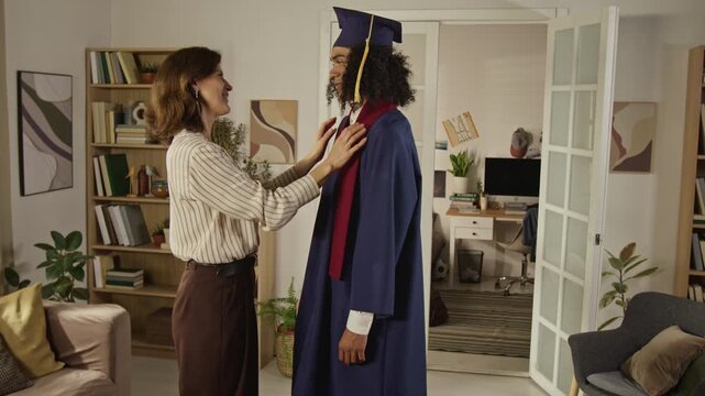 Side view shot of Caucasian middle aged mother adjusting graduation gown of Black teenager son at home. Footage is ideal for university enrollment campaigns and family support advertisements