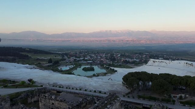 A drone flies forward, approaching the scenic natural lake situated below Pamukkale at dawn. The white travertine terraces and thermal waters reflect softly in the morning light, while dark mountains 