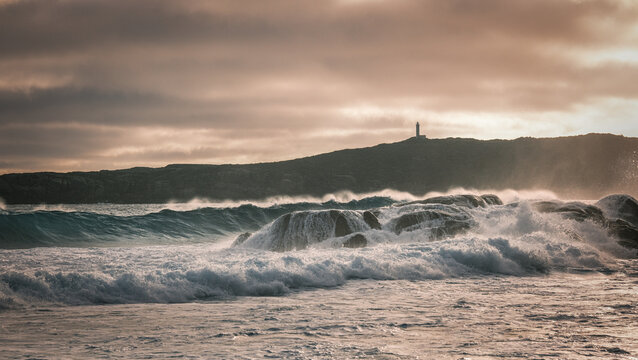 Aerial view of powerful ocean waves crashing against jagged coastal rocks with a distant lighthouse under a dramatic cloudy sky in Torndirrup, Western Australia, Australia.