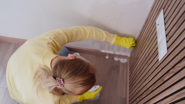 Top-down view of woman wearing protective yellow gloves and diligently cleaning moldy corner of wall with spray cleaner and sponge, removing fungus and mildew from home