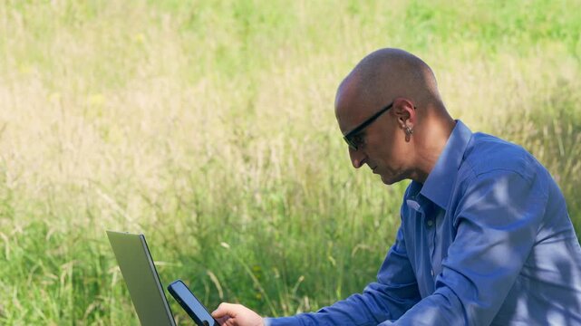 Professional man multi-tasking on a laptop and smartphone in a green meadow.
