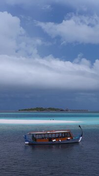 Aerial view of Dhigurah Island, Maldives, with a white sandbank, a dhoni gliding on navy to turquoise water, overwater bungalows, and storm clouds above.