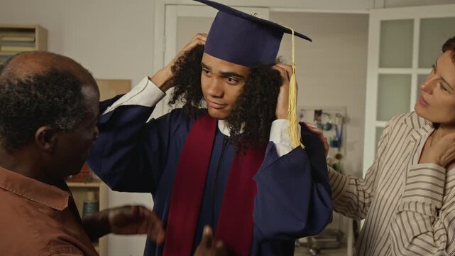 Handheld shot of African American teenager wearing graduation gown adjusting cap with help of Caucasian mother and Black father before hugging. Clip suits diverse family and higher education marketing