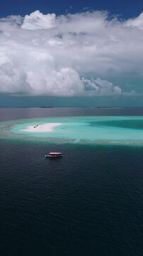 Aerial view of Dhigurah, Maldives, with a crescent sandbank, three beach tents, a drifting wooden dhoni, vivid turquoise lagoon, deep navy ocean, and cumulus clouds.