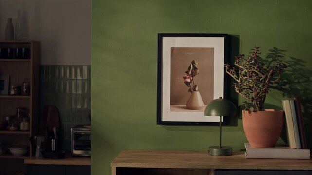 No people shot of green wall with framed art print above wooden table holding green lamp and potted plant. Footage serves as background for interior design promos or home decor marketing