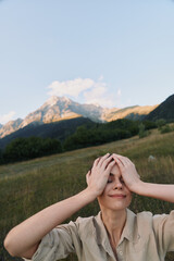 Fototapeta premium A woman outdoors in a mountain meadow holds her head with both hands, smiling softly. A moment of relief and self care in nature, capturing a lifestyle scene of calm, balance, and personal reflection.