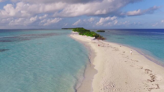 Aerial view of Dhigurah Island, Maldives shows a long sand spit, reef patches, shallow lagoon and deeper ocean, gentle waves, footprints, and people strolling at dusk