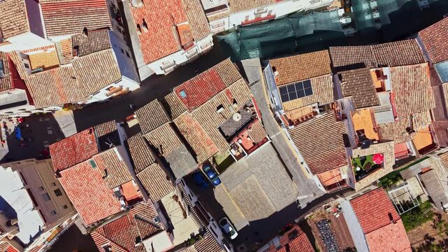 Aerial view of a small town showing closely packed houses with red roofs and narrow paths winding through the area. Cars are parked in the streets under bright sunlight.
