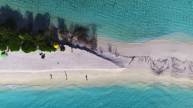 Aerial top down view of Dhigurah island, Maldives. Narrow sandbar, turquoise lagoon, deeper sea, green fringe, ripples, seaweed lines, umbrella, people strolling and wading.