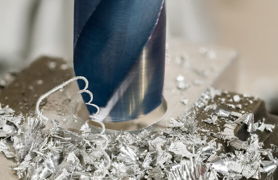 Closeup working solid core drill bit at enlarging hole in aluminum profile. Blue rotating steel cutting tool at drilling of workpiece clamped in bench vise jaws with shiny metal swarf and motion blur.