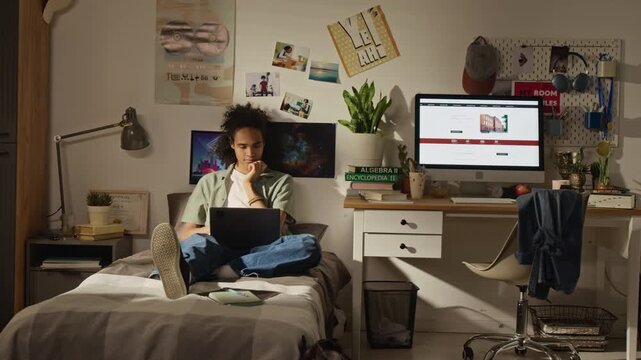 Full shot of African American teenager sitting on bed, using laptop for studying in bedroom. Clip is ideal for online education platforms, remote learning services, youth technology marketing