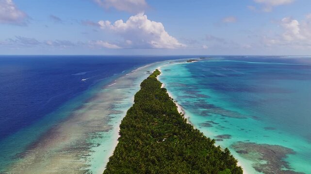 Aerial view of Dhigurah, Maldives, shows a narrow sandbar, palm canopy, deep navy ocean, turquoise lagoon, coral patches, and a small boat wake at midday