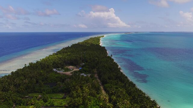Aerial view shows Dhigurah Island, Maldives, with fringing reef, pale sandbanks, white sand, coconut palms, lagoon and deep ocean as the shot slowly sweeps along