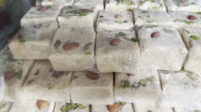 Close-up 4K footage of creamy Indian milk barfi squares topped with almonds and pistachios, neatly stacked behind a glass counter in a traditional sweet shop, highlighting festive dessert texture.