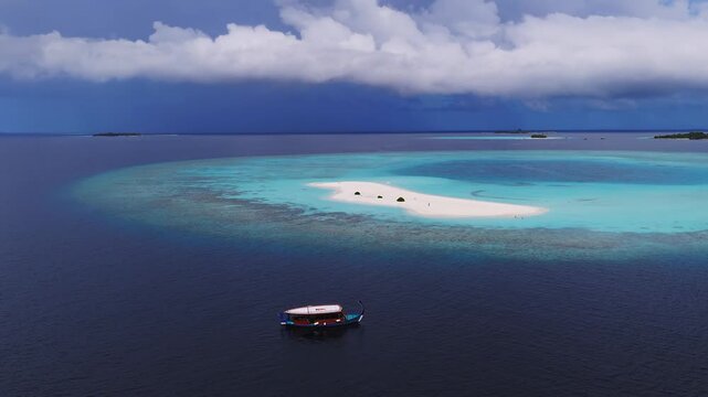 Aerial view of Dhigurah, Maldives shows a crescent sandbank, a drifting wooden dhoni, gentle waves on the reef, low clouds, and atolls on the distant horizon.