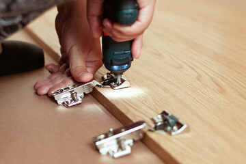 Fototapeta premium Close-up of a person using a power drill to attach metal hinges to a wooden cabinet door during home assembly, with a focus on the tools and materials involved