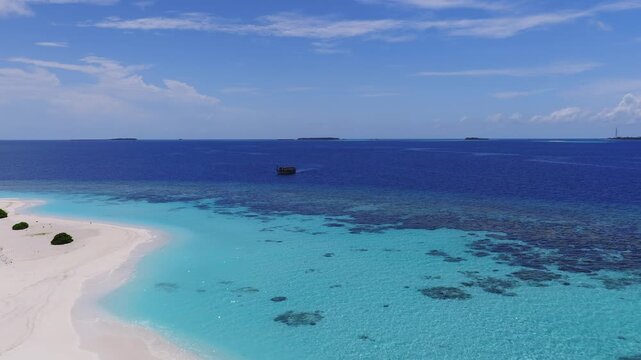 Aerial view of Dhigurah, Maldives, with crescent sand and turquoise lagoon, dark coral patches, a dhoni moving toward distant atolls and a faint tower at midday