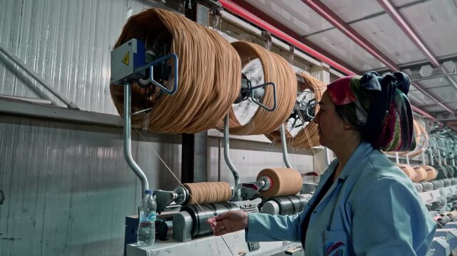 A female operator checks yarn from large bobbins on a winding machine in a textile factory. This video frame shows industrial yarn production, bobbin winding, quality control, and manufacturing proces
