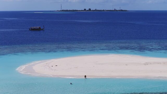 Aerial view of Dhigurah island, Maldives, with curved white sandbank, vivid turquoise lagoon, deep atoll waters, telecom towers, dhoni moving, walker and swimmer present