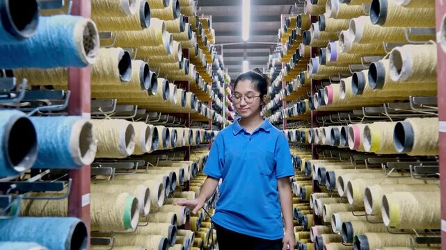 A female worker checks stock between shelves of yarn bobbins in a textile warehouse. This video frame shows yarn inventory, warehouse order, production preparation, and textile logistics.