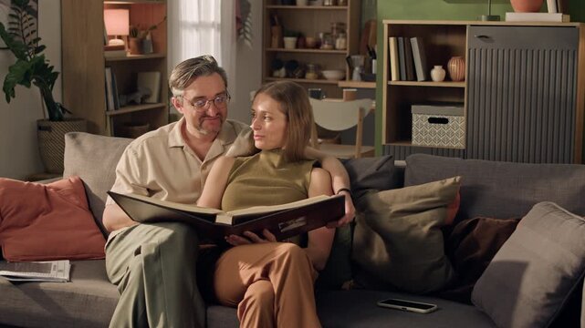 Wide shot of middle aged Caucasian man and young adult Caucasian woman sitting on sofa, flipping pages of photo album. Clip ideal for family heritage ads or memory preservation services