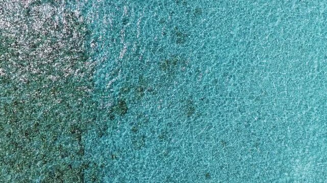 Aerial top down view over Dhigurah Island lagoon, Maldives. Turquoise water, coral reef patches, and sand textures shimmer in bright midday sun with gentle waves.