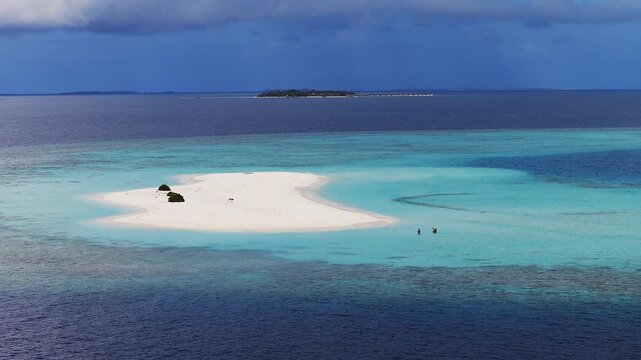 Aerial panoramic shows a crescent white sand cay off Dhigurah, Maldives. Two people wade in shallows, a small boat nears horizon, over water villas line distant island.