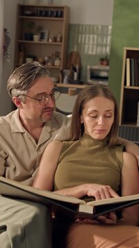 Vertical medium shot of middle aged Caucasian man and adult Caucasian woman sitting on sofa, turning pages of photo album. Clip perfect for family memory or photo book printing services