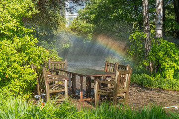 Naklejka premium A wooden patio set sits beneath cooling water mist. A colorful rainbow appears among lush trees and shrubs.
