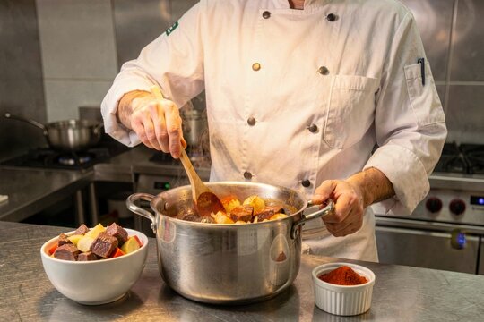 Chef in white uniform stirring Gulas Czech in large stainless steel pot. Modern kitchen with bright natural lighting, highlighting beef stew with paprika onions dumplings. Concept of culinary art.