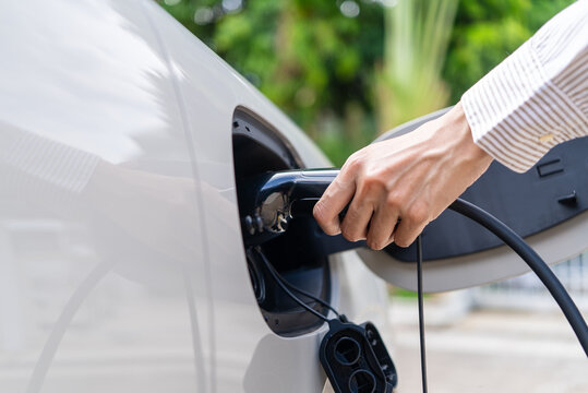 Close-up of an Asian woman's hand holding a charger plug connected to an electric car port. Concept of EV charging station alternative clean energy and sustainability.