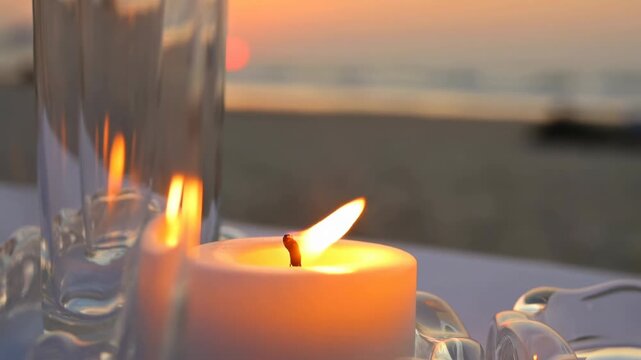 Candle flame tilting beside clear glass pieces at beach sunset with warm golden light, soft reflections, and calm romantic decor on textured tabletop near horizon