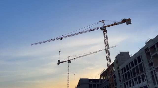 Tower crane lifting heavy equipment at construction site. Building structure development at sunset. Engineering machinery working on urban infrastructure project.