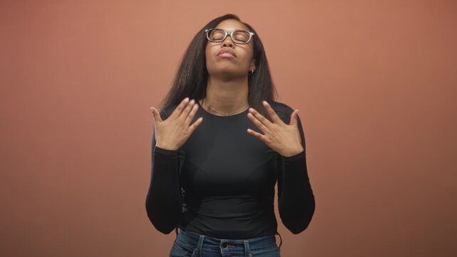 Woman with glasses and jeans gesturing with hands and touching chest, eyes closed, framed against a seamless studio backdrop; quiet contemplation.