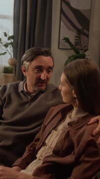 Vertical medium shot of middle aged Caucasian couple sitting on sofa, talking, and embracing. Footage suitable for relationship counseling or mental health marketing campaigns