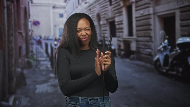 Woman young african american holding a hair straightener with one hand, grimacing and waving the other hand near face on a narrow street in a city; playful amusement.