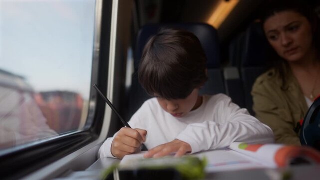 Young boy studying during train journey with mother nearby, everyday family travel scene highlighting portable learning, concentration, education and productive use of travel time