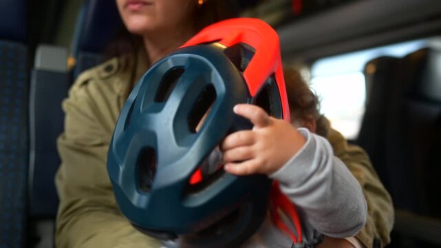 Toddler trying to put on bicycle helmet while sitting on mother lap during family train journey, playful travel moment highlighting curiosity, learning, safety awareness and family connection