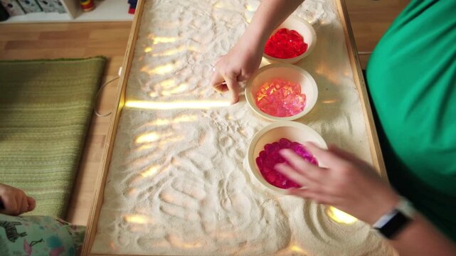 Woman creating patterns for her sensory sand art display. Creative learning process with glowing light box, bowls of colorful gems and tactile kinetic sand play.