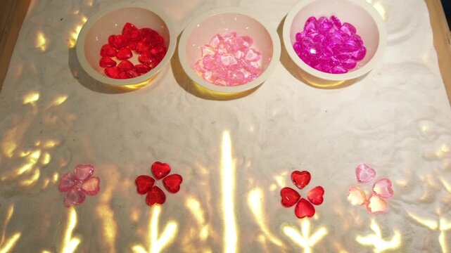 Bowls filled with small heart shaped gems placed on a illuminated sand table. Decorative flower patterns made of glass hearts for Valentine activity or art design.