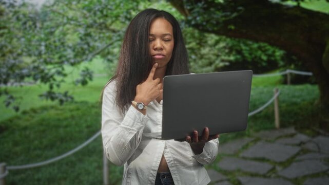 Woman holding laptop with finger on chin, studying the screen beside a paved path in a forest; thoughtful learning.