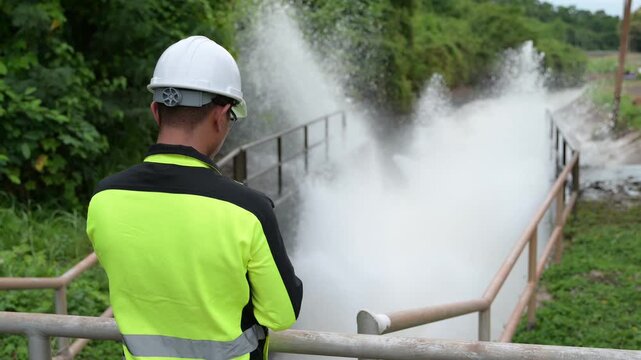 Professional male environmental engineer wearing a hard hat inspecting rushing water at a dam spillway while taking notes on a clipboard for public infrastructure safety and flood control.