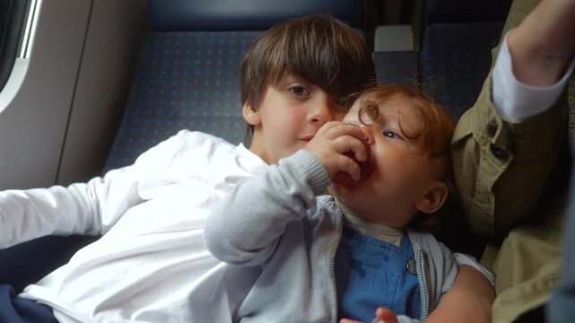 Toddler eating fresh nectarine while older brother kisses his cheek during passenger train journey, heartwarming family travel moment highlighting sibling affection and childhood bonding