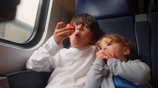 Toddler eating fresh nectarine while relaxing beside older sibling during passenger train journey, authentic family travel moment highlighting healthy nutrition and childhood comfort