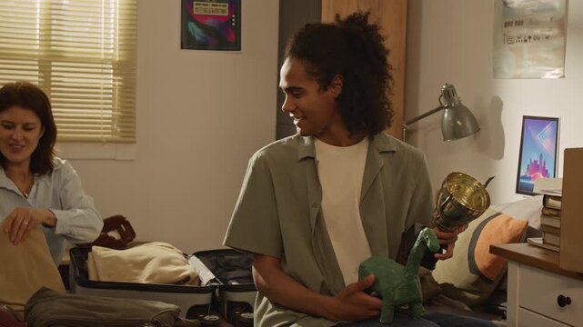 Handheld shot of Black teenager holding dinosaur toy and placing trophy into cardboard box while Caucasian middle-aged mother packs suitcase. For relocation services or college transition campaigns