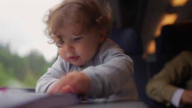 Curious toddler looking through illustrated storybook while riding passenger train with mother, authentic family travel moment highlighting early childhood learning and exploration