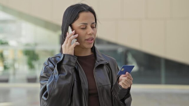 Woman young hispanic holding smartphone to ear while checking a creditcard in a building entrance; concerned payment issue.