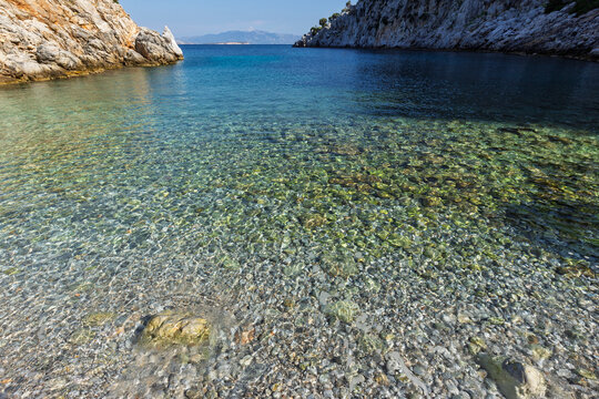 Small pebbles and stones under shallow water by a beach in a cove surrounded by rugged and hilly coastline by the Aegean Sea on Kalymnos island in Greece on a sunny day.